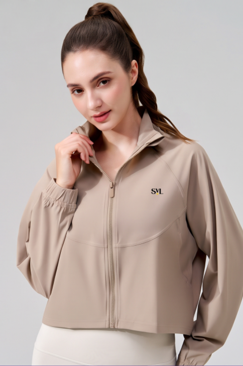A woman wearing a cropped, long-sleeve athletic jacket in a light taupe-beige color. The jacket features a mock neck, a front zipper, elastic cuffs, and a small black 'SVL' logo on the left chest.