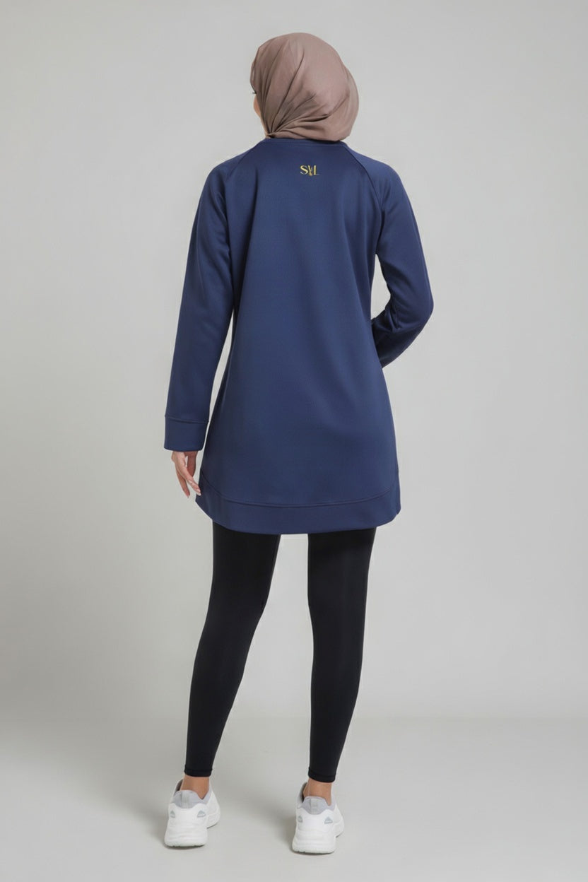 Rear view of a female player wearing a navy blue SVL modest padel tunic, showing the gold embroidered logo on the upper back and a taupe hijab.