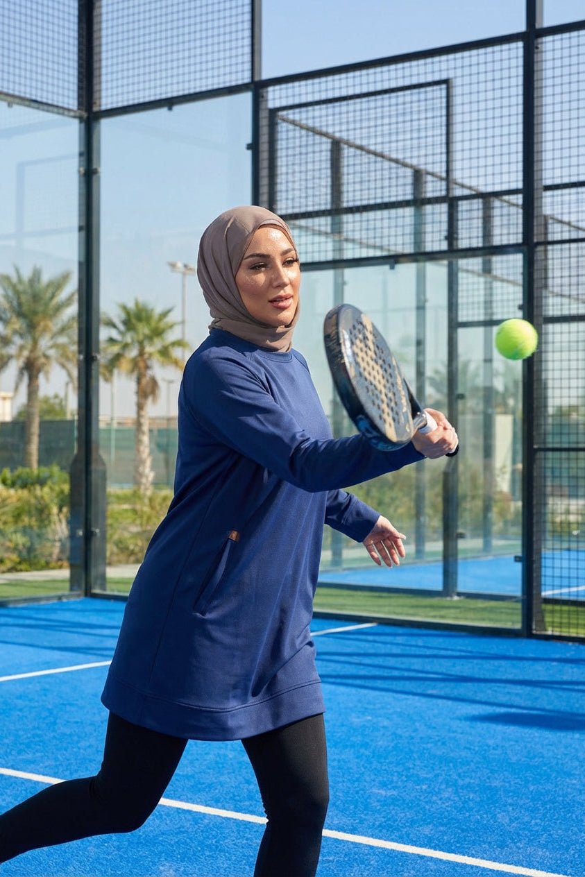 A female player on a blue padel court mid-swing, wearing a modest navy blue SVL tunic and a taupe hijab.