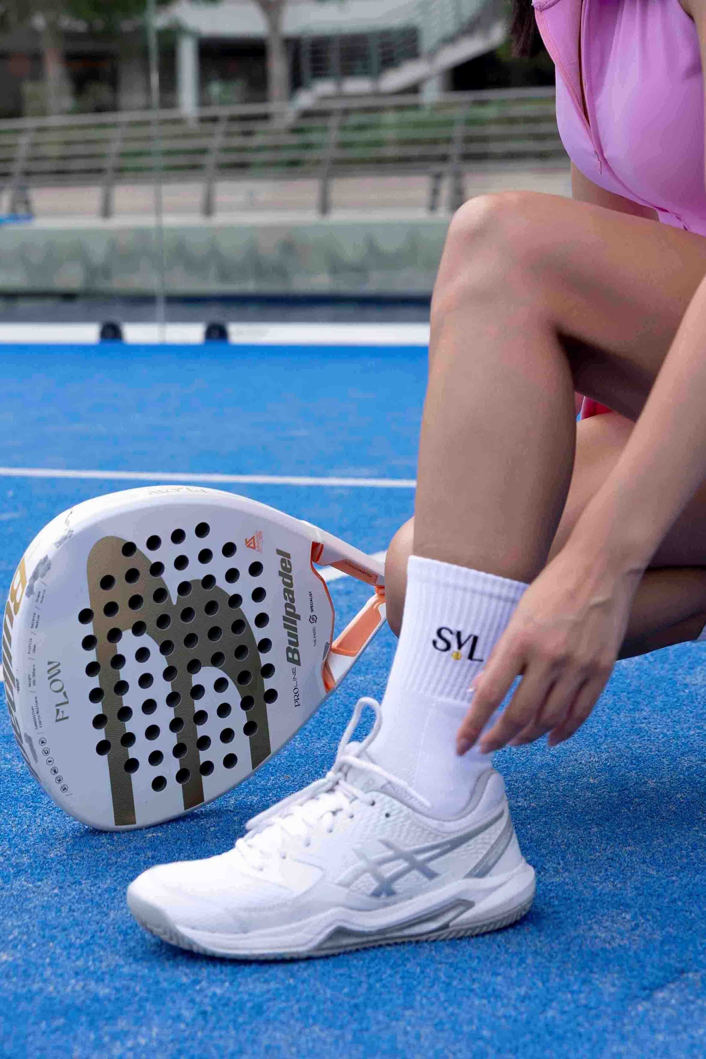 Lifestyle shot of a player adjusting their white SVL grip socks on a blue padel court, showing the embroidered logo and non-slip sole design.
