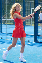A female padel player in motion on a blue court wearing the red SVL Kate skirt and matching zip-polo top.