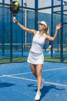 A female padel player competing on a blue padel court wearing the white SVL Kate skirt and matching zip-up polo top.
