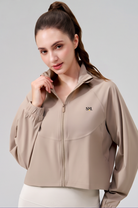 A woman wearing a cropped, long-sleeve athletic jacket in a light taupe-beige color. The jacket features a mock neck, a front zipper, elastic cuffs, and a small black 'SVL' logo on the left chest.