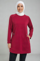 A female player wearing a modest maroon SVL padel tunic with long sleeves, side pockets, and a white hijab, designed for comfort and performance on the court.