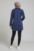 Rear view of a female player wearing a navy blue SVL modest padel tunic, showing the gold embroidered logo on the upper back and a taupe hijab.